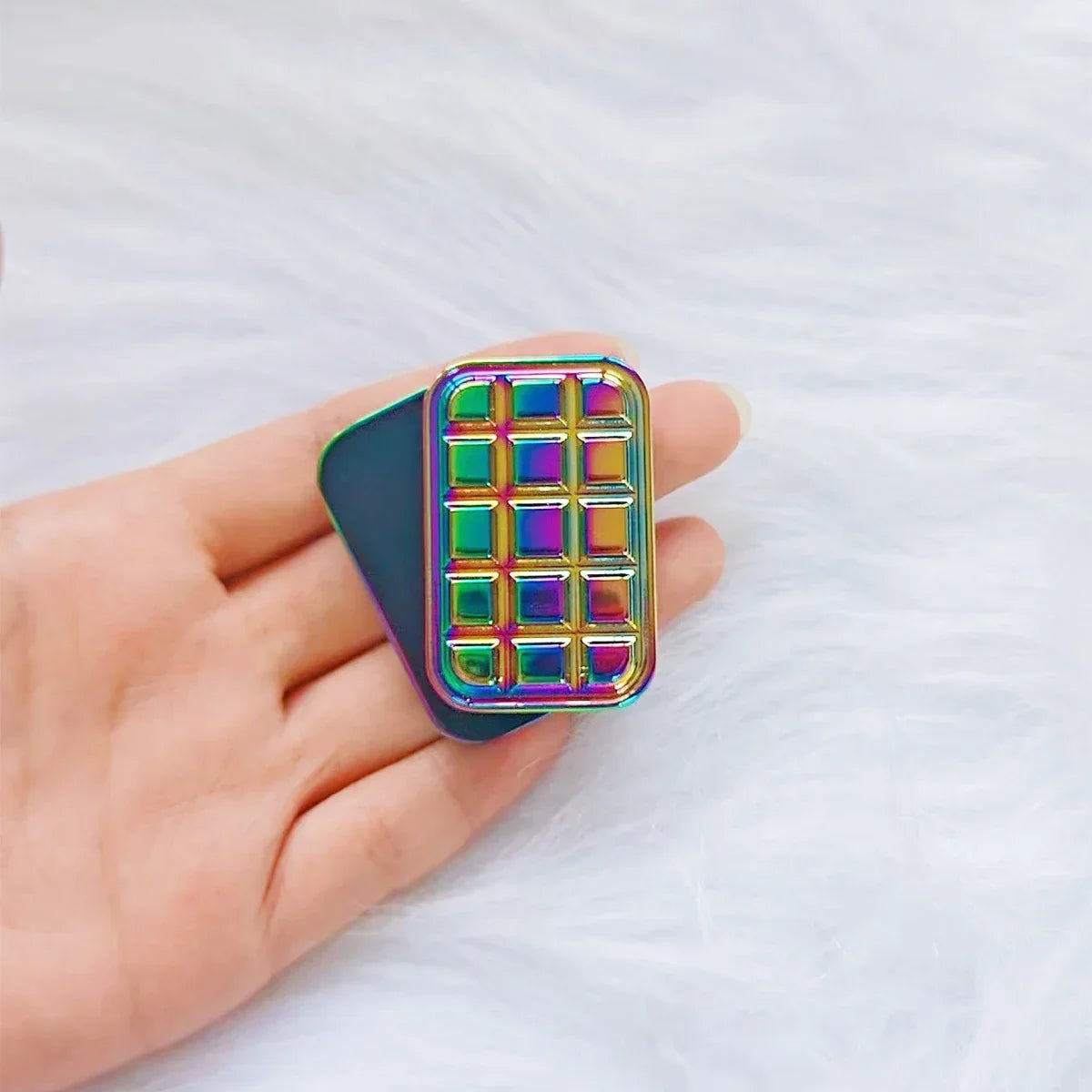 Hand holding a rainbow metallic fidget toy with a grid pattern on a soft white background